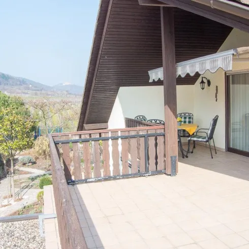 Ein Balkon mit Blick auf eine grüne Landschaft und Berge im Hintergrund. Tische und Stühle laden zum Entspannen ein.