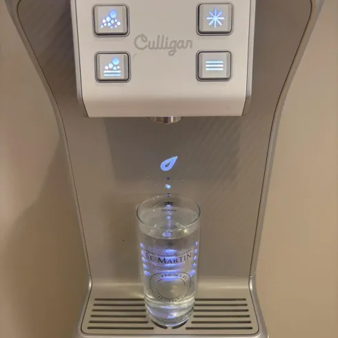 Ein moderner Wasserspender mit leuchtenden Tasten und einem Glas darunter. Das Glas ist teilweise mit klarem Wasser gefüllt.