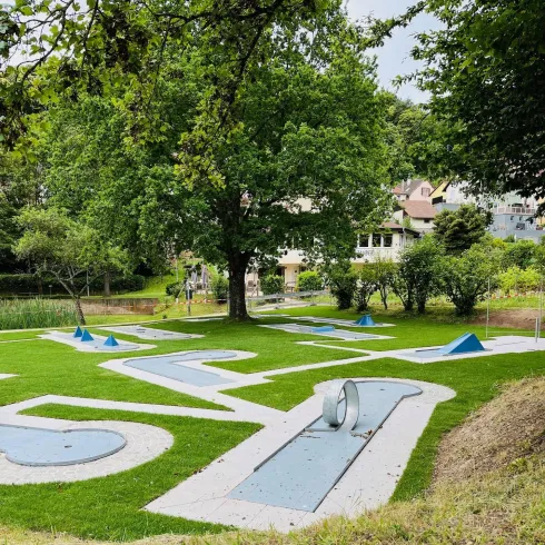Eine Minigolfanlage in einem Park mit grünem Rasen und großen Bäumen. Im Hintergrund sind Wohnhäuser sichtbar.