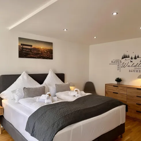 Ein modernes Schlafzimmer mit einem Doppelbett, stilvollen Kissen und einer warmen Holzkommode. An der Wand hängt ein Bild einer Stadtlandschaft sowie inspirierende Zitate über die Natur.