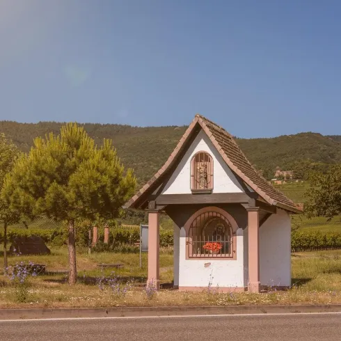 Eine kleine Kapelle steht an einem ruhigen Landweg, umgeben von Bäumen und Weinbergen. Der Himmel ist klar und sonnig.