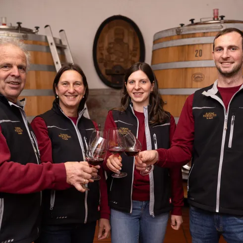 Eine Gruppe von vier Personen in einem Weinkeller hält Gläser mit Rotwein. Im Hintergrund sind große Holzfässer zu sehen.