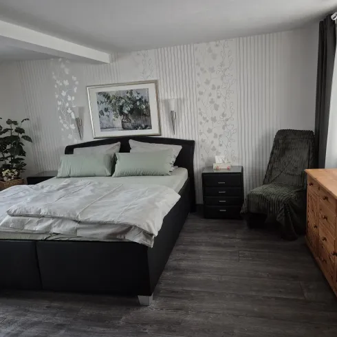 Ein modernes Schlafzimmer mit einem großen, komfortablen Bett und hellen Kissen. Daneben befindet sich eine gemütliche Sitzecke und eine Holzkommode.