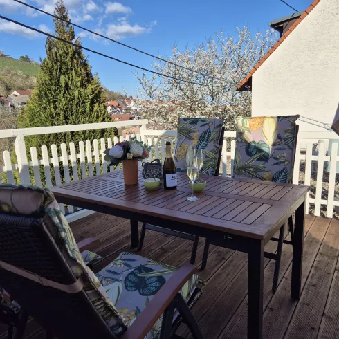 Ein einladender Balkon mit einem Holztisch und zwei Stühlen. Auf dem Tisch stehen Blumen und ein Flasche Wein, umgeben von einer schönen Landschaft.