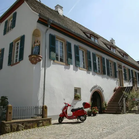Ein charmantes Gebäude mit weißen Wänden und grünen Fensterläden. Vor dem Haus steht ein rotes Motorrad und eine angelegte Treppe führt zum Eingangsbereich.