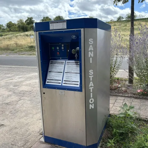 Eine Sani-Station mit einem blauen Gehäuse steht auf einem Bürgersteig. Im Hintergrund sind Bäume und ein leicht bewölkter Himmel zu sehen.