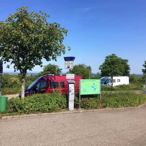 Ein Parkplatz mit einem roten Fahrzeug und einem Wohnmobil. Im Hintergrund sind Bäume und ein blauer Himmel zu sehen.