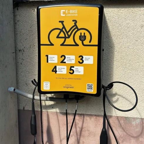 Eine E-Bike-Ladestation an einer Wand. Sie ist gelb und hat mehrere Ladeanschlüsse sowie Anweisungen für die Nutzung.