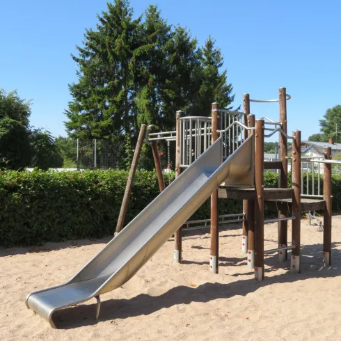 Ein Spielplatz mit einer Rutsche aus Metall und Holzspielgeräten. Im Hintergrund sind grüne Sträucher und Bäume zu sehen.