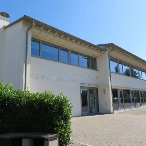 Ein modernes Gebäude mit großen Fenstern und einem klaren, blauen Himmel. Im Vordergrund sind grüne Sträucher zu sehen.