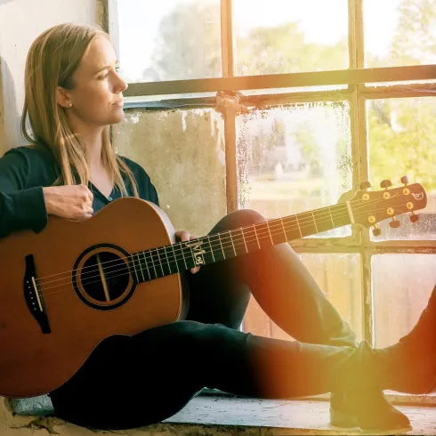 Eine Frau sitzt am Fenster und spielt Gitarre. Die natürliche Beleuchtung schafft eine warme und inspirierende Atmosphäre.
