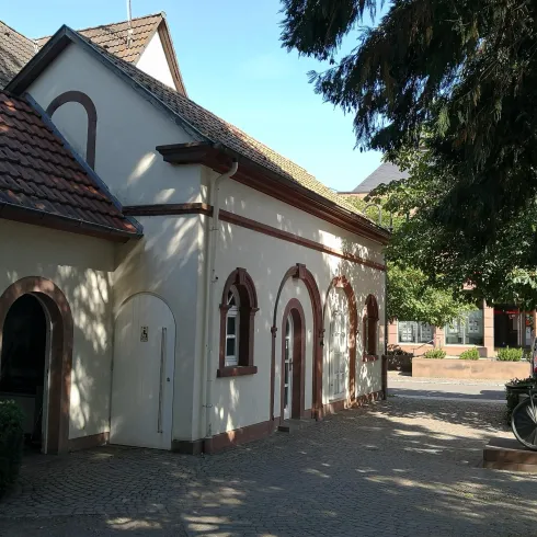 Ein charmantes Gebäude mit weißen Wänden und roten Akzenten. Bäume spenden Schatten und der Himmel ist blau und klar.
