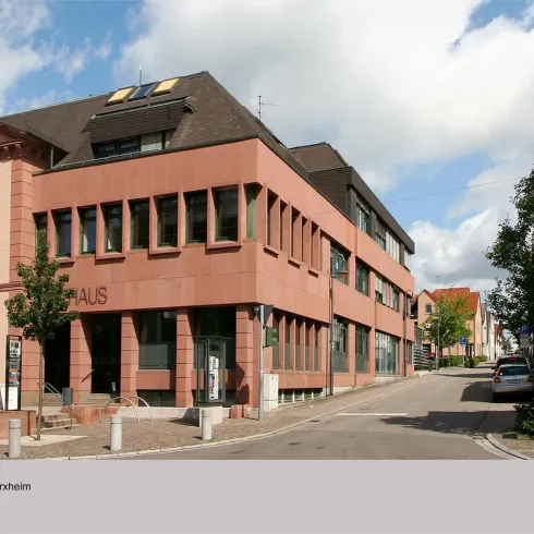 Ein modernes Gebäude aus roten Ziegeln steht an einer ruhigen Straße. Rechts sieht man ein traditionelles Fachwerkhaus.