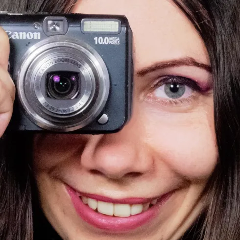 Eine Frau hält eine Kamera vor ihr Gesicht und lächelt. Sie hat lange, dunkle Haare und trägt einen farbigen Lidschatten.