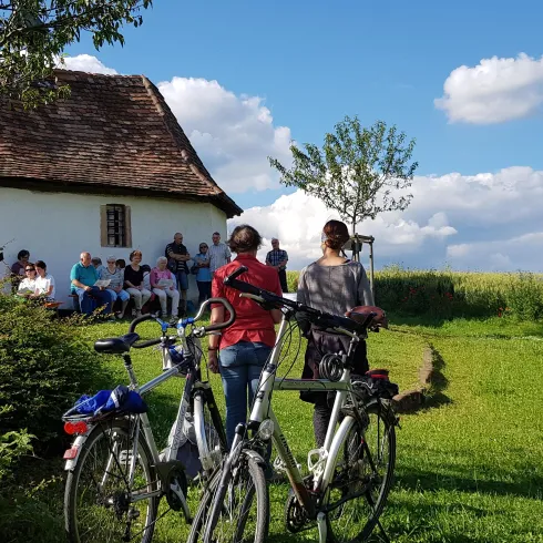 Zwei Personen stehen mit Fahrrädern auf einer Wiese vor einer Kapelle. Im Hintergrund sitzen mehrere Menschen und genießen die Landschaft.