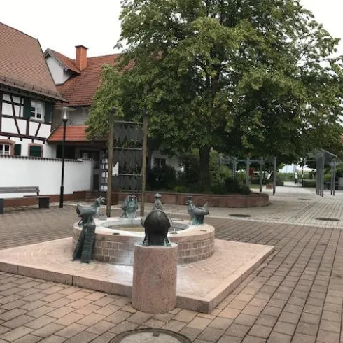 Ein hübscher Platz mit einem Brunnen in der Mitte und mehreren Bänken. Umgeben von Bäumen und einem traditionellen Gebäude.