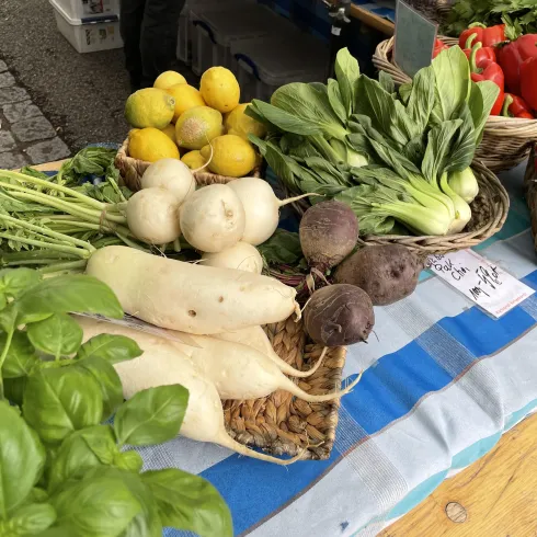Ein frischer Marktstand mit einer Auswahl an Gemüse und Obst. Darunter sind Radieschen, Paprika, Zwiebeln und Zitrone zu sehen.