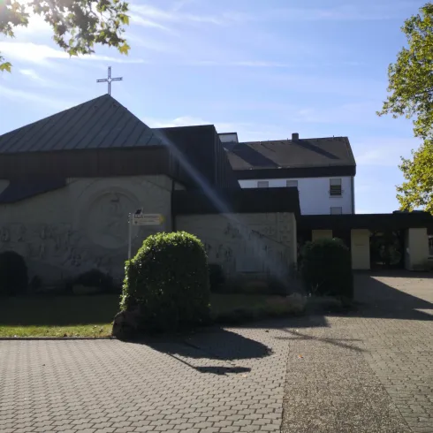 Ein Gebäude mit einem grauen Putz und einem abgedunkelten Dach. Im Vordergrund sind die gepflasterte Auffahrt und einige Sträucher zu sehen.