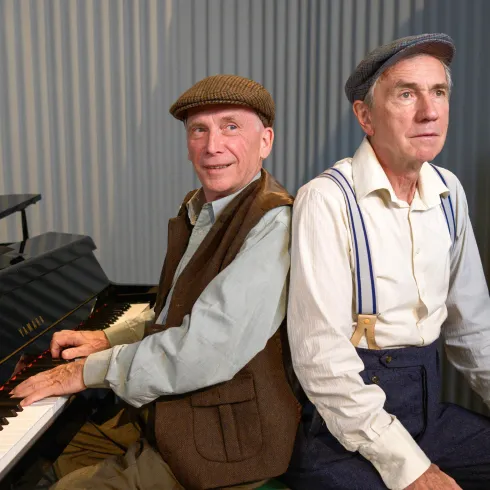 Zwei ältere Männer sitzen Rücken an Rücken, während einer am Klavier spielt. Beide tragen traditionelle Kleidung und wirken freundlich.