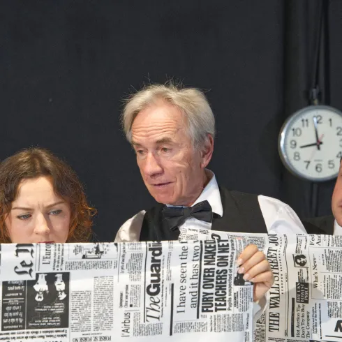 Drei Personen betrachten intensiv ein großes Stück Zeitungspapier. Im Hintergrund steht eine Uhr, die die Zeit anzeigt.
