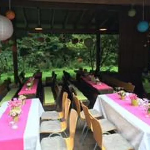 Ein festlich dekorierter Veranstaltungsraum mit langen Tischen. Die Tische sind mit bunten Tüchern und Blumen geschmückt, umgeben von grüner Natur.