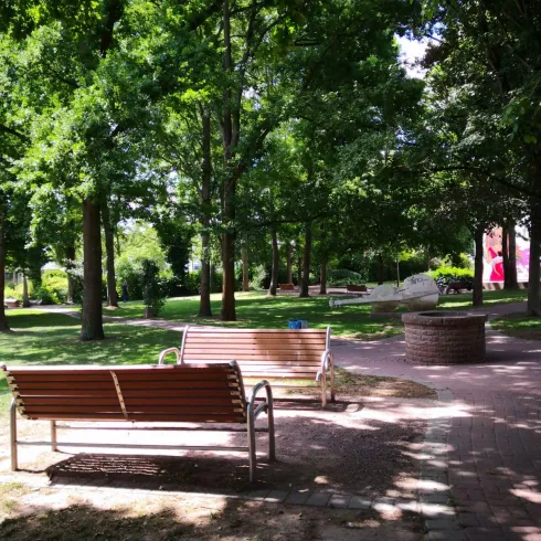 Ein ruhiger Park mit grünen Bäumen und Bänken. Der Weg ist gepflastert und lädt zum Entspannen ein.