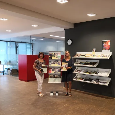 Zwei Frauen stehen in einem modernen Raum neben einem Informationsständer. Im Hintergrund sind Regale mit verschiedenen Materialien und ein Empfangsbereich zu sehen.