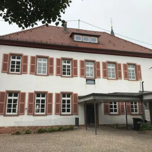 Ein historisches Gebäude mit roter Fensterverkleidung und weißer Fassade. Der Eingang ist überdacht und es gibt eine Pflanzfläche im Vordergrund.