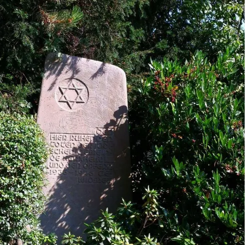 Ein Denkmal mit einem Davidstern steht umgeben von grünen Sträuchern. Die Inschrift ist teilweise verdeckt, aber gut sichtbar.