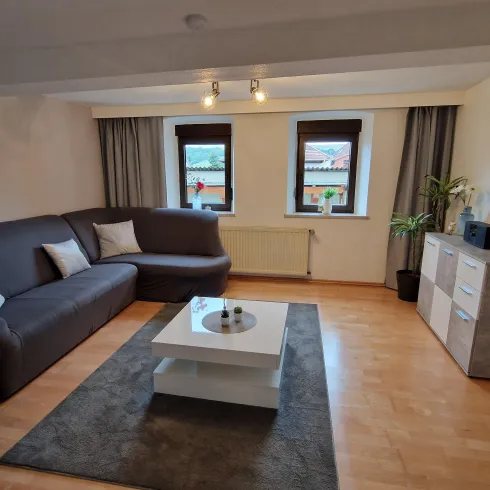 Ein modernes Wohnzimmer mit einer grauen Couch und einem Couchtisch. Große Fenster lassen viel Licht herein, und es gibt Pflanzen und Dekorationen im Raum.
