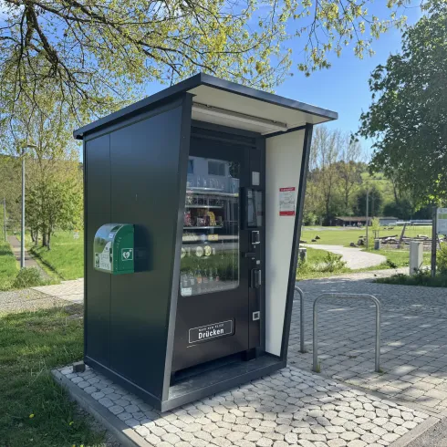 Ein moderner Verkaufsautomat in einer kleinen Kabine. Die Umgebung besteht aus saftigem Grün und ist einladend gestaltet.