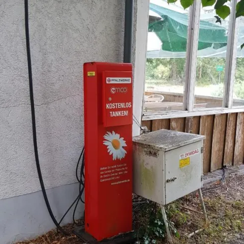 Eine rote Säule mit der Aufschrift „Kostenlos Tanken“ steht neben einem geschlossenen Gebäude. Daneben befindet sich eine kleine metallene Box.