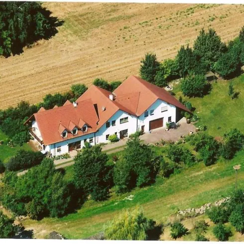Ein großes Haus mit rotem Dach, umgeben von Bäumen und Grünflächen. Im Hintergrund sind landwirtschaftliche Felder zu sehen.