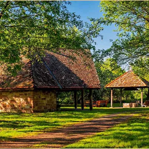 Ein idyllisches Gebäude aus Stein mit einem schrägen Dach inmitten vieler Bäume. Nebenan steht ein überdachter Sitzbereich auf einer Wiese.