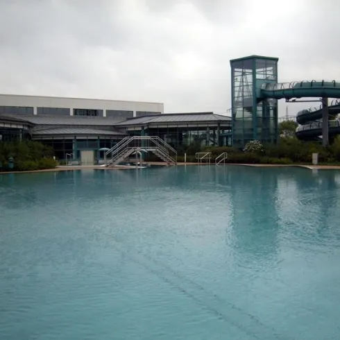 Ein großes Schwimmbad mit glattem Wasser und umliegenden Pflanzen. Im Hintergrund befindet sich ein Gebäude mit einer Rutsche.