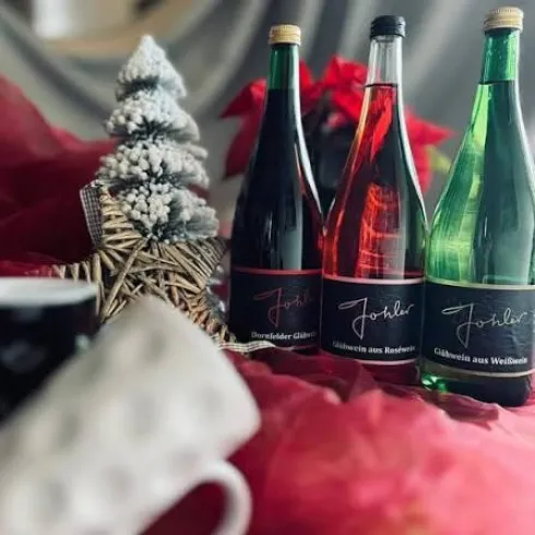 Drei Flaschen Glühwein in verschiedenen Farben stehen vor einer festlichen Dekoration. Im Hintergrund sind ein Weihnachtsbaum und rote Pflanzen zu sehen.