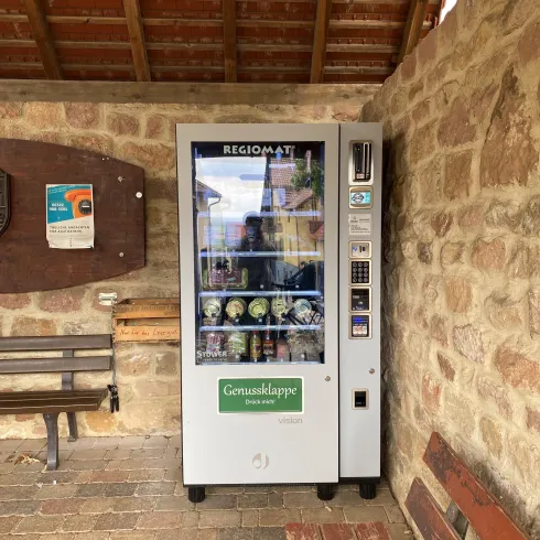 Ein Automat mit regionalen Produkten steht in einem überdachten Bereich. Er enthält verschiedene Flaschen und Dosen.