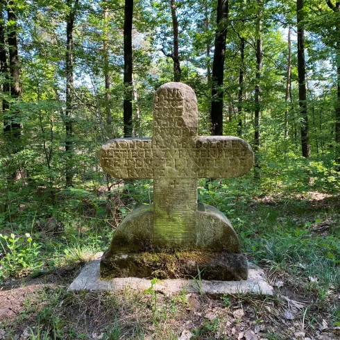 Ein altes, steinernes Kreuz steht im Wald, umgeben von Bäumen und Sträuchern. Die Inschriften auf dem Kreuz sind nur teilweise lesbar.