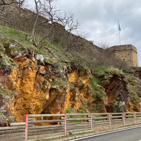 Eine steile Felswand mit orangefarbenen Ablagerungen und grünem Bewuchs. Im Hintergrund ist eine alte Mauer mit einem Fahnenmast zu sehen.