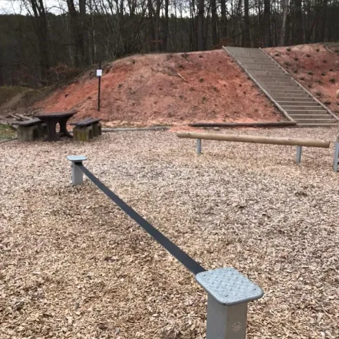 Ein Spielplatz mit einer langen Balancestange und Sitzmöglichkeiten aus Holz. Im Hintergrund sind Treppen und ein kleiner Hügel zu sehen.