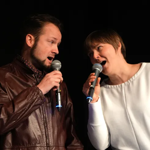 Ein Mann und eine Frau stehen auf der Bühne und singen miteinander in ein Mikrofon. Beide haben Spaß und sind konzentriert auf ihre Darbietung.