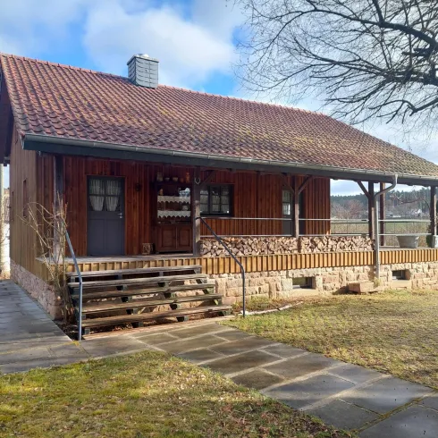 Ein hübsches Holzhaus mit einer Veranda und einem roten Dach. Die Umgebung ist grün und einladend, ideal für entspannte Tage im Freien.