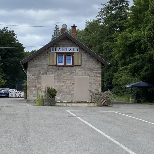 Ein kleines, steinernes Gebäude mit einem Schild, das "Der Wirt" zeigt. Es steht in einem leeren Parkplatz, umgeben von Bäumen und grünem Gelände.