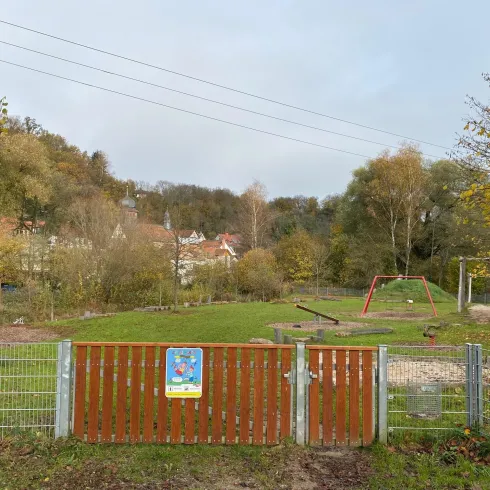 Ein Spielplatz mit einer grünen Wiese und einigen Spielgeräten. Der Bereich ist mit einem Zaun und einem Tor umgeben, umgeben von Bäumen und Hügeln.