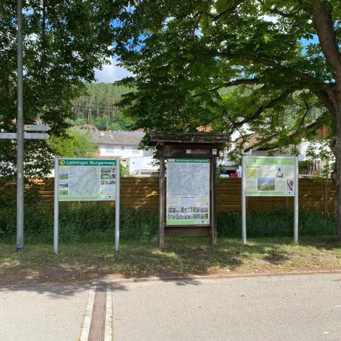 Eine Informationsstelle mit mehreren Tafeln steht unter einem großen Baum. Im Hintergrund sind Gebäude und eine bewaldete Landschaft sichtbar.