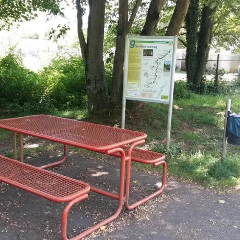 Ein roter Picknicktisch mit Bänken steht unter schattigen Bäumen. Neben dem Tisch befindet sich ein Informationsschild und ein Mülleimer.
