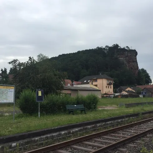 Eine ruhige Bahnhofslandschaft mit einem steilen Felsen im Hintergrund. Im Vordergrund sind Gleise und einige Gebäude zu sehen.