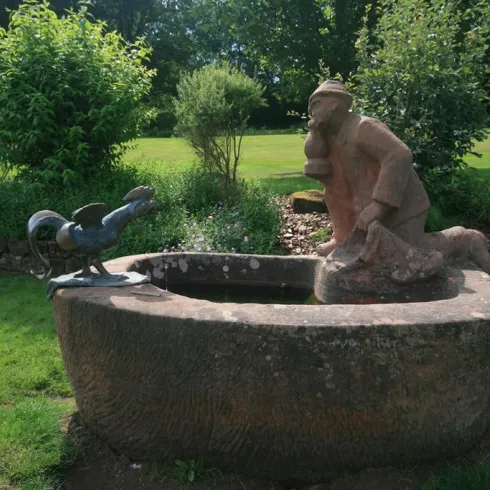 Eine Statue eines Mannes, der an einem Brunnen sitzt, während ein Hahn ihm gegenüber steht. Umgeben von grünem Rasen und Sträuchern.
