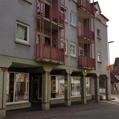Ein Gebäude mit mehreren Stockwerken und Balkonen in einer Stadt. Im Hintergrund sind weitere traditionelle Häuser zu sehen.