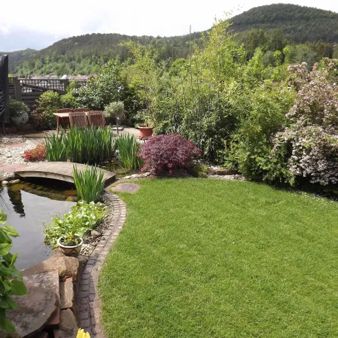 Ein schöner Garten mit grünem Rasen, Blumen und einem kleinen Teich. Im Hintergrund sind Hügel und Bäume zu sehen.